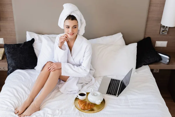 Vista ad alto angolo di giovane donna in accappatoio seduta vicino al computer portatile e vassoio per la colazione sul letto — Foto stock