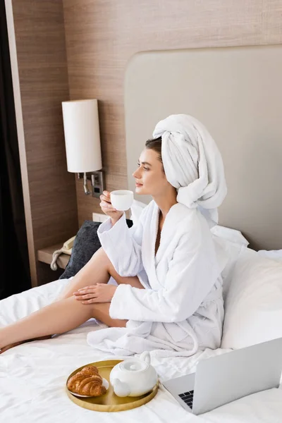 Jeune femme en peignoir tenant tasse de café près de l'ordinateur portable et plateau avec petit déjeuner — Photo de stock