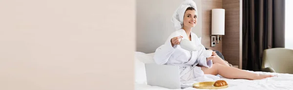 Panoramic crop of young woman in bathrobe holding cup and teapot near laptop and tray with breakfast — Stock Photo