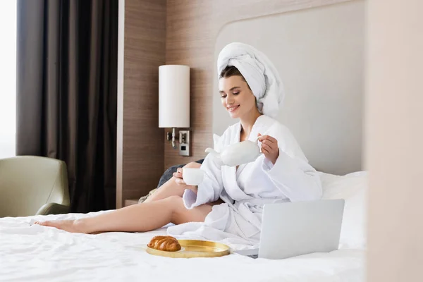 Woman in bathrobe holding cup and teapot near laptop and tray with breakfast on bed — Stock Photo