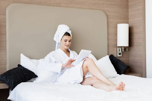 Mujer en toalla y albornoz blanco mirando la tableta digital en la habitación del hotel - foto de stock