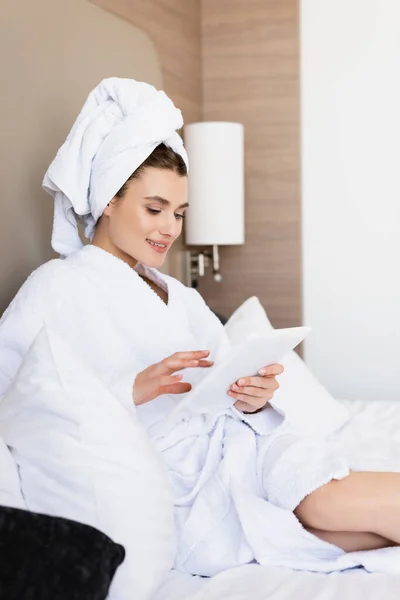Mujer joven en toalla y albornoz blanco mirando la tableta digital en la habitación del hotel - foto de stock