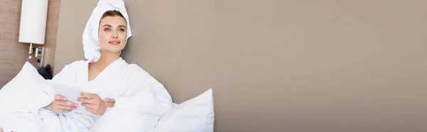 Imagen horizontal de mujer joven en toalla y albornoz blanco sosteniendo tableta digital en habitación de hotel - foto de stock