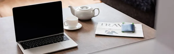 Coltura panoramica di laptop con schermo bianco vicino teiera, tazza, giornale di viaggio e passaporto con biglietto aereo sul tavolo — Foto stock