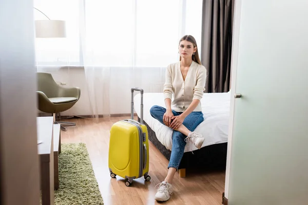 Selective focus of young woman in jeans sitting on bed near with yellow luggage in hotel room — Stock Photo