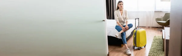 Concepto horizontal de mujer joven en jeans sentados en la cama cerca con equipaje amarillo en la habitación de hotel - foto de stock