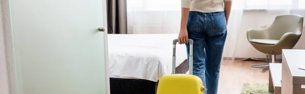Panoramic crop of young woman in jeans walking with yellow luggage in hotel room — Stock Photo