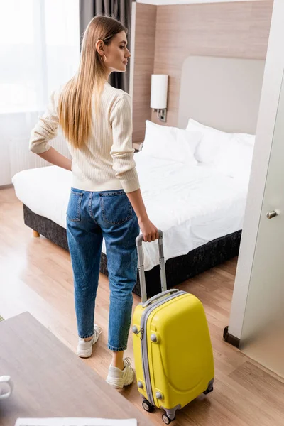 Young woman in jeans standing with yellow luggage in hotel room — Stock Photo