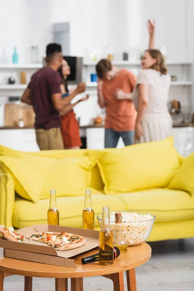 Foco seletivo de pizza e cerveja na mesa perto de sofá amarelo e amigos multiétnicos dançando no fundo — Fotografia de Stock