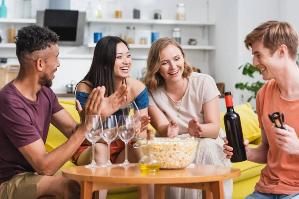 Jovem homem segurando garrafa de vinho tinto perto de amigos multiculturais animado — Fotografia de Stock