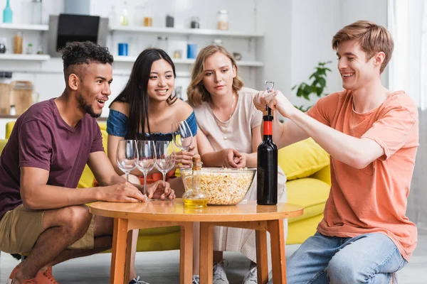 Giovane aprendo bottiglia di vino rosso vicino gioiosi amici multiculturali — Foto stock