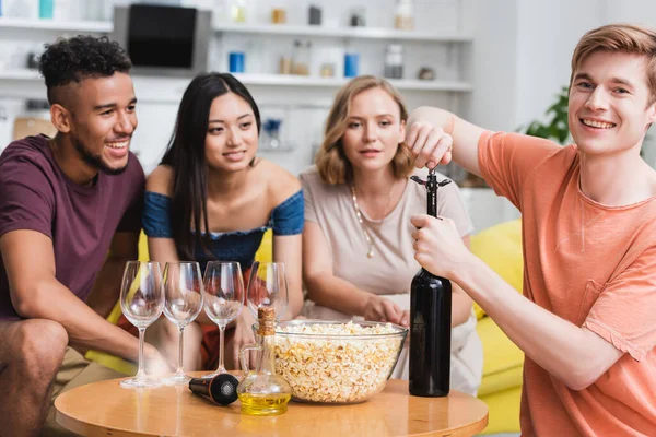 Jovem abrindo garrafa de vinho tinto perto de amigos multiculturais — Fotografia de Stock