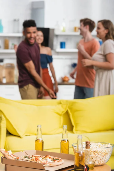 Selective focus of beer and pizza on table near yellow sofa and multiethnic friends dancing on background — Stock Photo