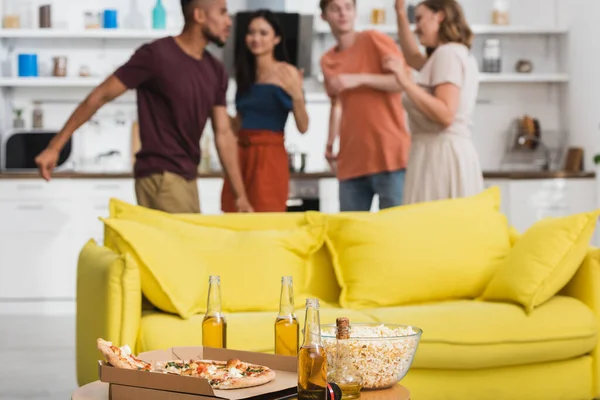 Foco seletivo de cerveja e pizza na mesa perto de sofá amarelo e amigos multiculturais dançando no fundo — Fotografia de Stock