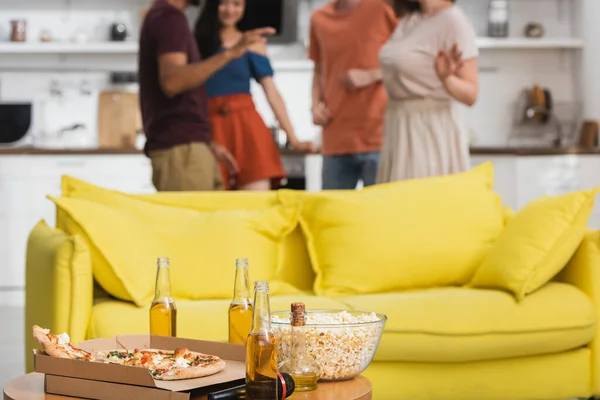 Selective focus of pizza and beer on table near yellow sofa and multicultural friends dancing on background — Stock Photo