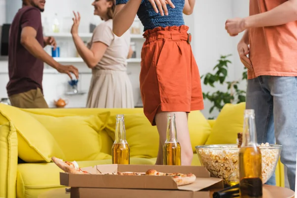 Selective focus of pizza and beer on table near yellow sofa and multiethnic friends having fun on background — Stock Photo