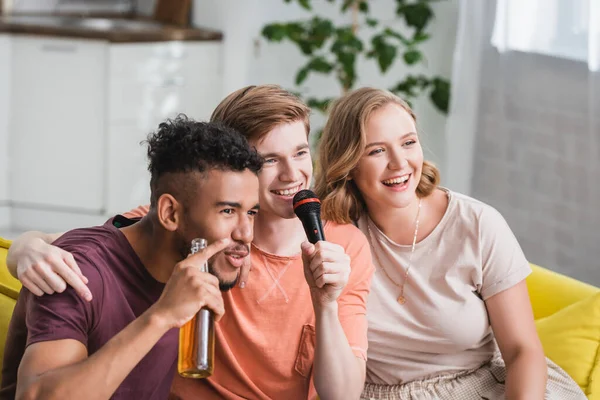 Africano americano hombre celebración botella de cerveza cerca multicultural amigos cantando karaoke - foto de stock