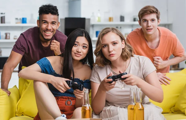 KYIV, UCRANIA - 28 de julio de 2020: alegre hombre afroamericano mostrando un gesto ganador cerca de mujeres multiculturales jugando videojuegos - foto de stock