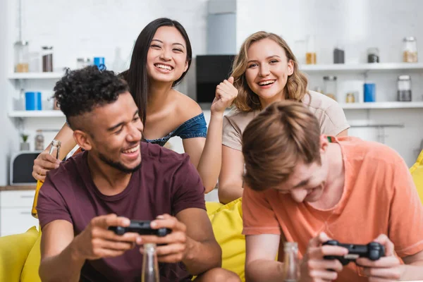 KYIV, UCRANIA - 28 de julio de 2020: enfoque selectivo de la mujer asiática emocionada mostrando el gesto ganador cerca de amigos multiculturales jugando videojuegos - foto de stock
