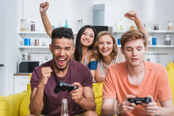 KYIV, UKRAINE - JULY 28, 2020: selective focus of excited women showing winner gesture near multicultural friends playing video game — Stock Photo