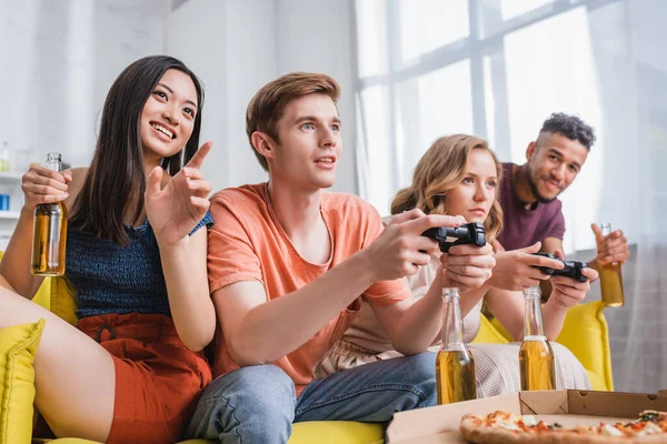 KYIV, UKRAINE - JULY 28, 2020: excited asian woman holding beer and pointing with finger near friends playing video game — Stock Photo