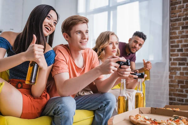 KYIV, UKRAINE - JULY 28, 2020: excited asian woman holding beer and showing thumb up near friends playing video game — Stock Photo