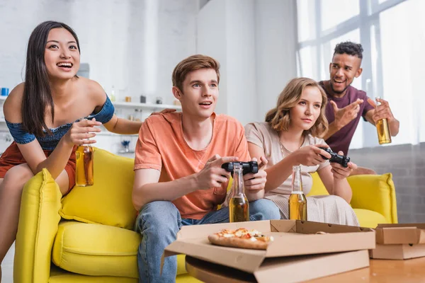 KYIV, UKRAINE - JULY 28, 2020: excited asian woman and african american man holding bottles of beer near friends playing video game — Stock Photo