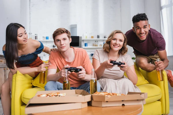 KYIV, UKRAINE - JULY 28, 2020: african american man and asian woman holding bottles of beer near friends playing video game — Stock Photo