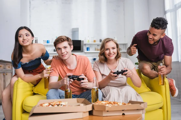 KYIV, UKRAINE - JULY 28, 2020: african american man holding bottle of beer near multiethnic friends playing video game — Stock Photo