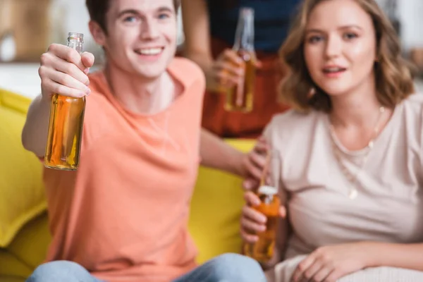 Избирательный фокус молодого человека и женщины, держащих бутылки пива и смотрящих в камеру во время вечеринки — стоковое фото