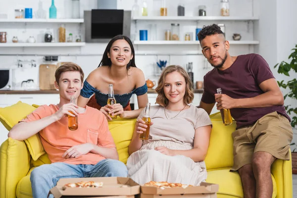 Joyful multiethnic friends with bottles of beer watching tv near pizza during party — Stock Photo