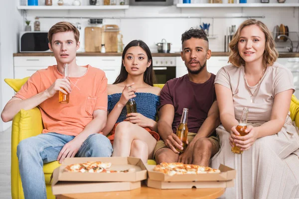 Multicultural friends with bottles of beer watching tv near pizza during party — Stock Photo