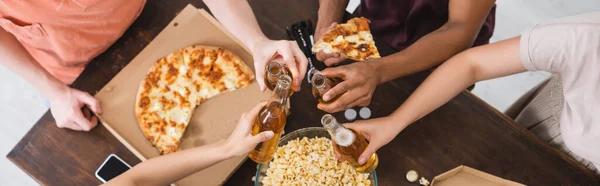 Vista ritagliata di amici multiculturali clinking bottiglie di birra vicino pizza durante la festa, intestazione del sito web — Foto stock