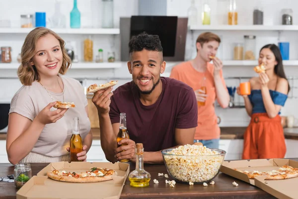 Zufriedene multiethnische Freunde, die während einer Party Pizza essen, in die Kamera schauen — Stockfoto