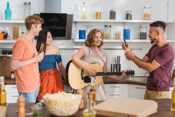 Mujer joven tocando la guitarra para amigos multiculturales, mientras que el hombre afroamericano tomando fotos en el teléfono inteligente - foto de stock