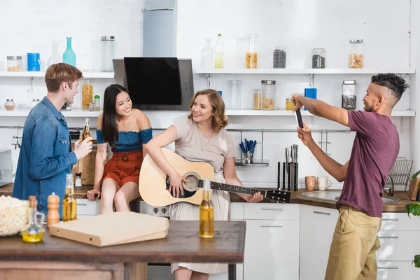 Africano americano homem tirar foto no smartphone enquanto jovem mulher tocando guitarra para amigos multiculturais — Fotografia de Stock