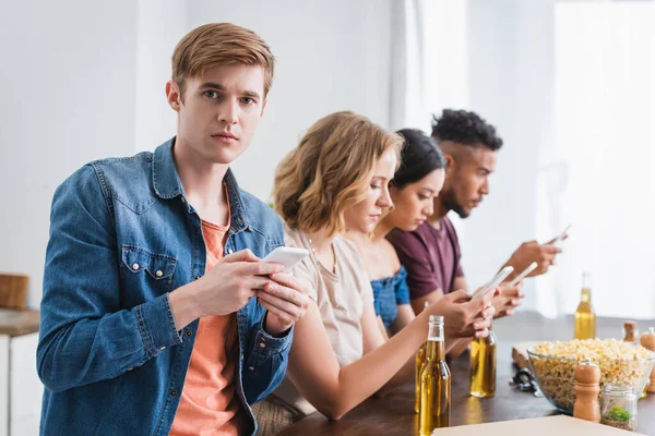 Селективное внимание мультикультурных друзей, общающихся на мобильных телефонах возле пива и попкорна во время вечеринки — стоковое фото