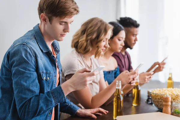 Selective focus of multiethnic friends messaging on smartphones near beer during party — Stock Photo