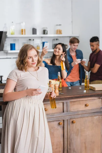 Вибірковий фокус молодої жінки, що тримає пиво і смартфон біля багатоетнічних друзів на фоні — стокове фото