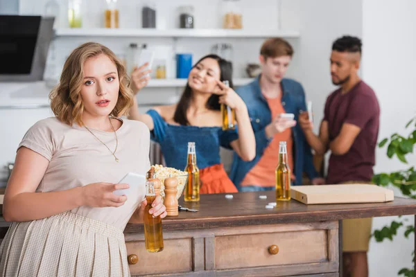 Selective focus of young woman with beer and smartphone looking at camera near multicultural friends on background — Stock Photo