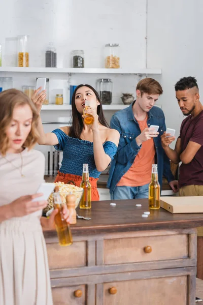 Selektiver Fokus einer asiatischen Frau, die Bier trinkt und Selfies mit multiethnischen Freunden macht, die während einer Party auf Mobiltelefonen chatten — Stockfoto