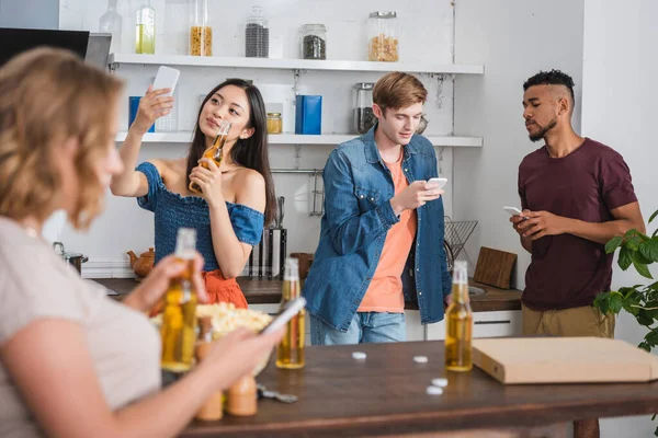 Selektiver Fokus einer asiatischen Frau mit Bier und Selfie in der Nähe multikultureller Freunde mit Smartphones während einer Party — Stockfoto