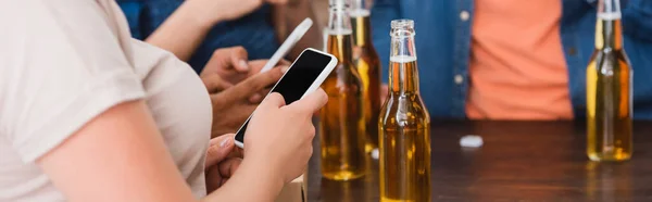 Vista parcial de amigos multiétnicos charlando en teléfonos inteligentes cerca de la cerveza durante la fiesta, cosecha horizontal - foto de stock