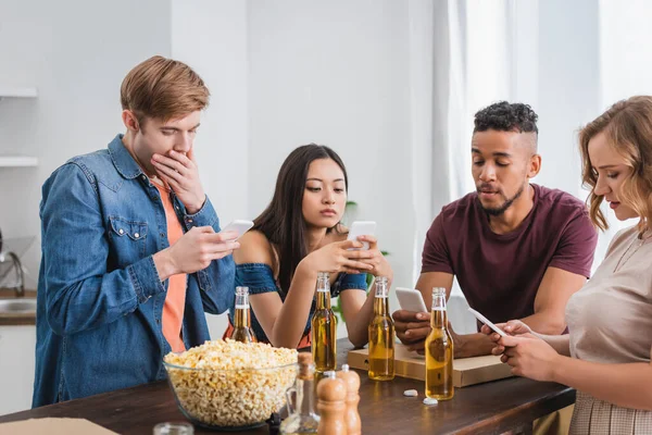 Enfoque selectivo de amigos multiétnicos utilizando teléfonos móviles cerca de cerveza y palomitas de maíz durante la fiesta - foto de stock