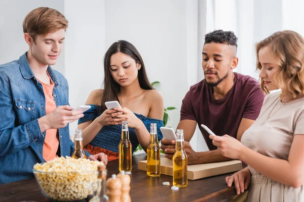 Selective focus of multiethnic friends messaging on smartphones near beer and popcorn during party — Stock Photo