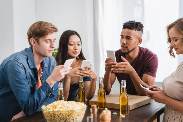 Foco seletivo de amigos multiculturais conversando em smartphones perto de cerveja e pipoca durante a festa — Fotografia de Stock