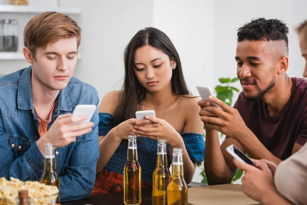 Selektiver Fokus multikultureller Freunde, die während einer Party in der Nähe von Bier auf Smartphones chatten — Stockfoto