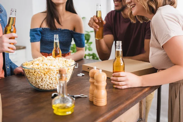 Cropped view of multiethnic friends near bowl of popcorn and bottles of beer — Stock Photo