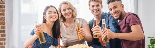 Conceito panorâmico de amigos multiétnicos alegres olhando para a câmera enquanto segurando garrafas de cerveja — Fotografia de Stock