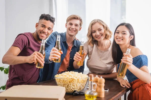 Selektiver Fokus fröhlicher multiethnischer Freunde, die in die Kamera schauen, während sie Bierflaschen in der Hand halten — Stockfoto